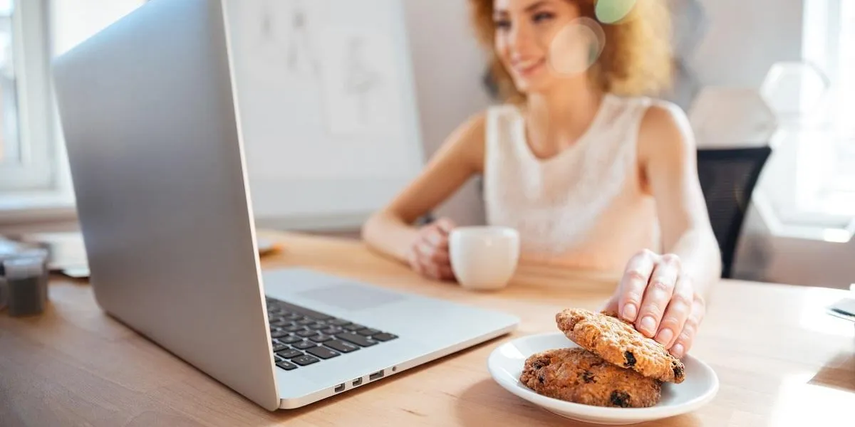 smiling pretty redhead young business woman drinking coffee with cookies on workplace and using laptop smiling pretty redhead young business woman drinking coffee with cookies on workplace and using laptop